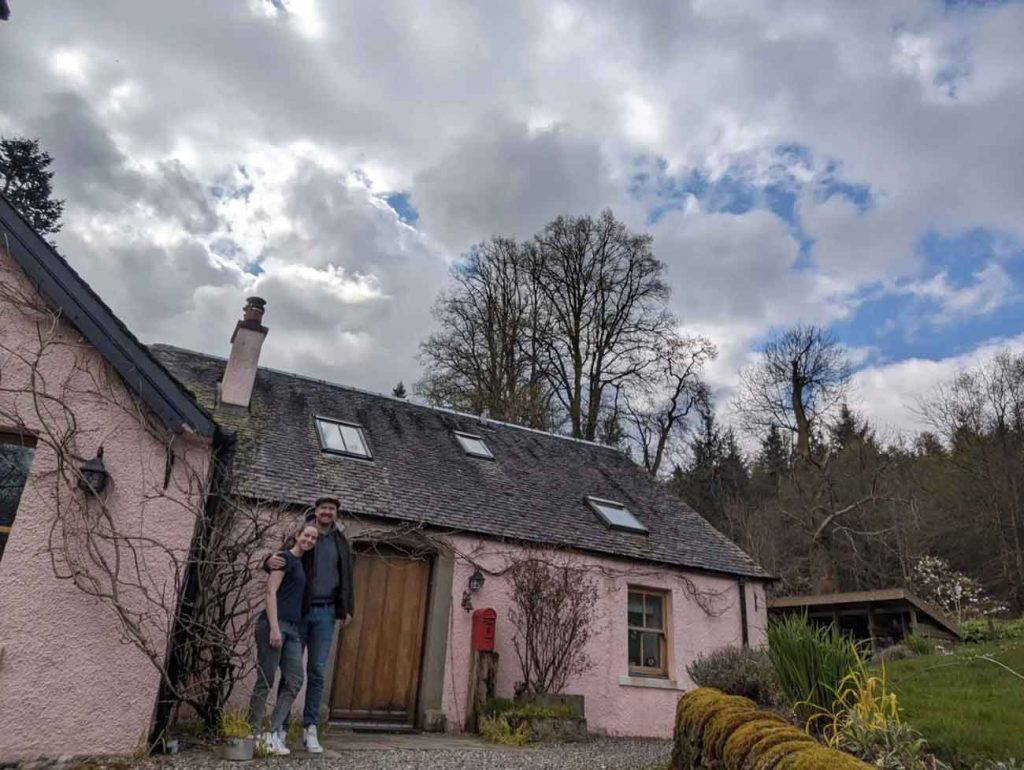 Couple standing outside a pink countryside cottage under a cloudy sky