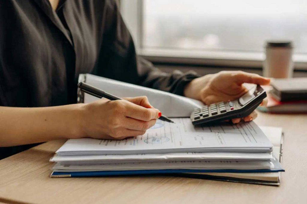 Person using calculator and writing on documents at a desk near a window